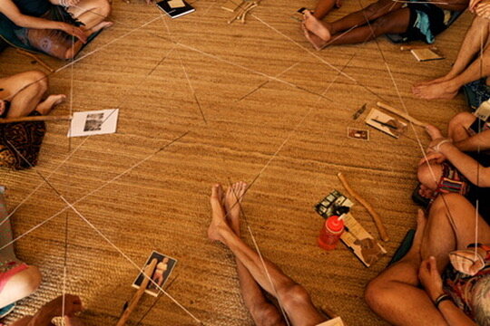 Overhead view of a group circle exercise — yarn strung between participants creating a web of relational connection on the floor.